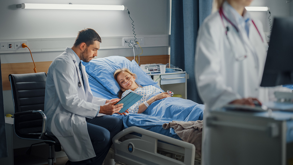 Frau im Krankenhausbett wird von einem Arzt betreut, der neben ihr auf einem Stuhl sitzt als Symbolbild für die gesetzliche Krankenversicherung